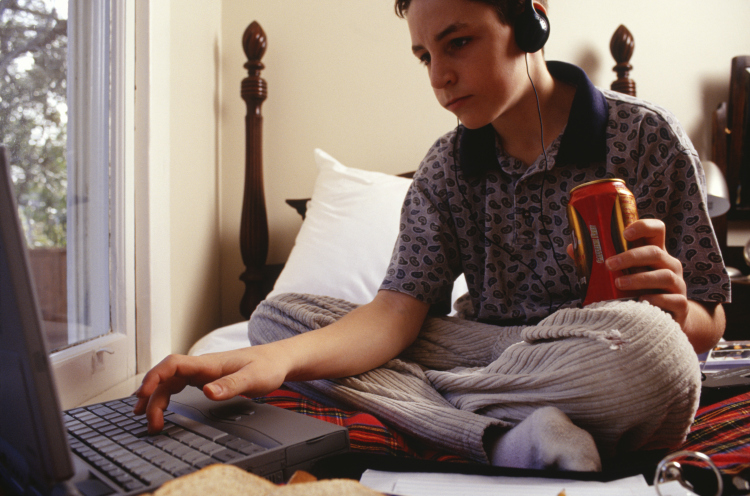 Teenage boy drinking energy drink, using laptop, sitting on bed